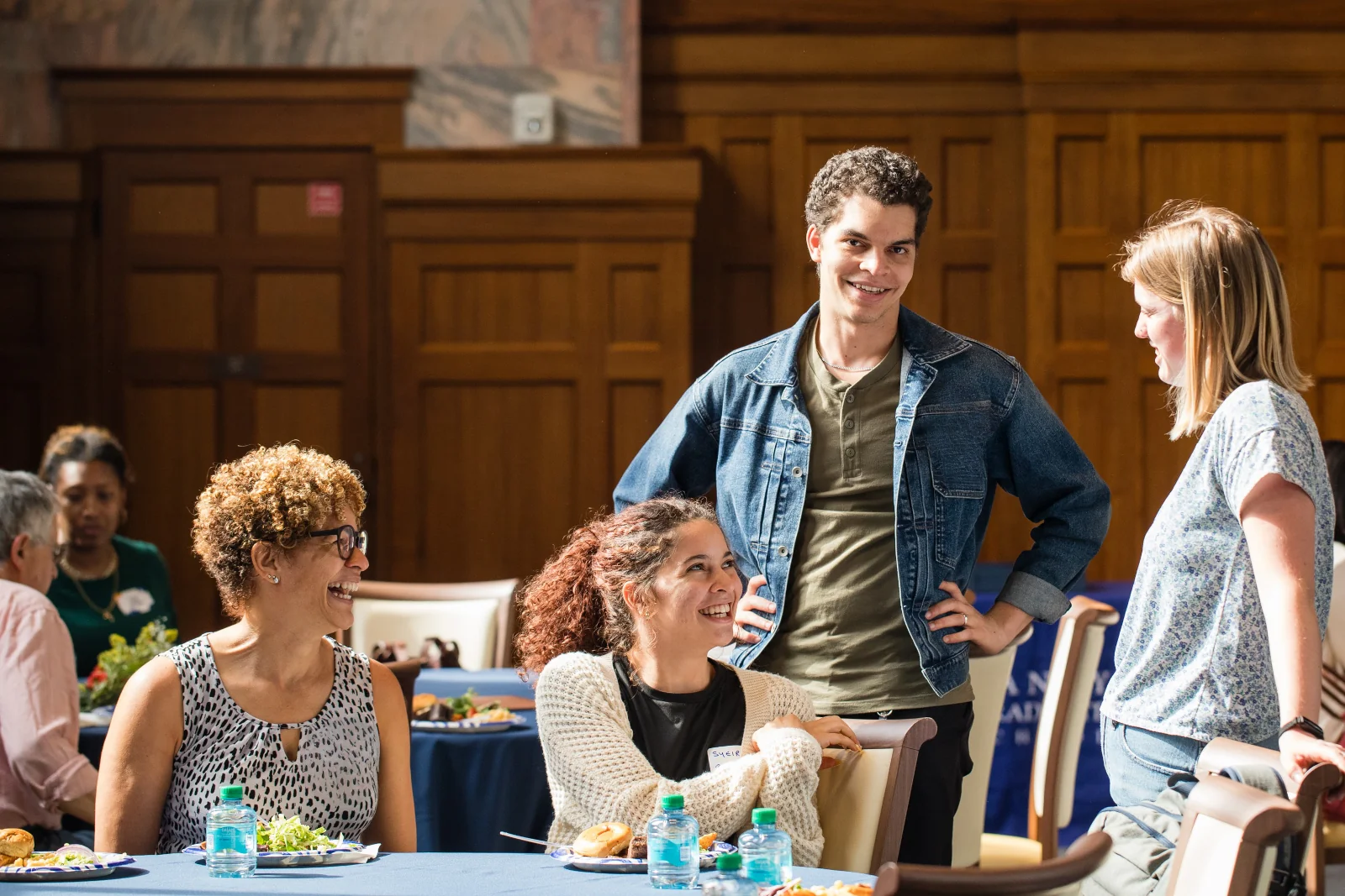 Four students laugh and talk together at a round table during a social event in a wood-paneled campus space.
