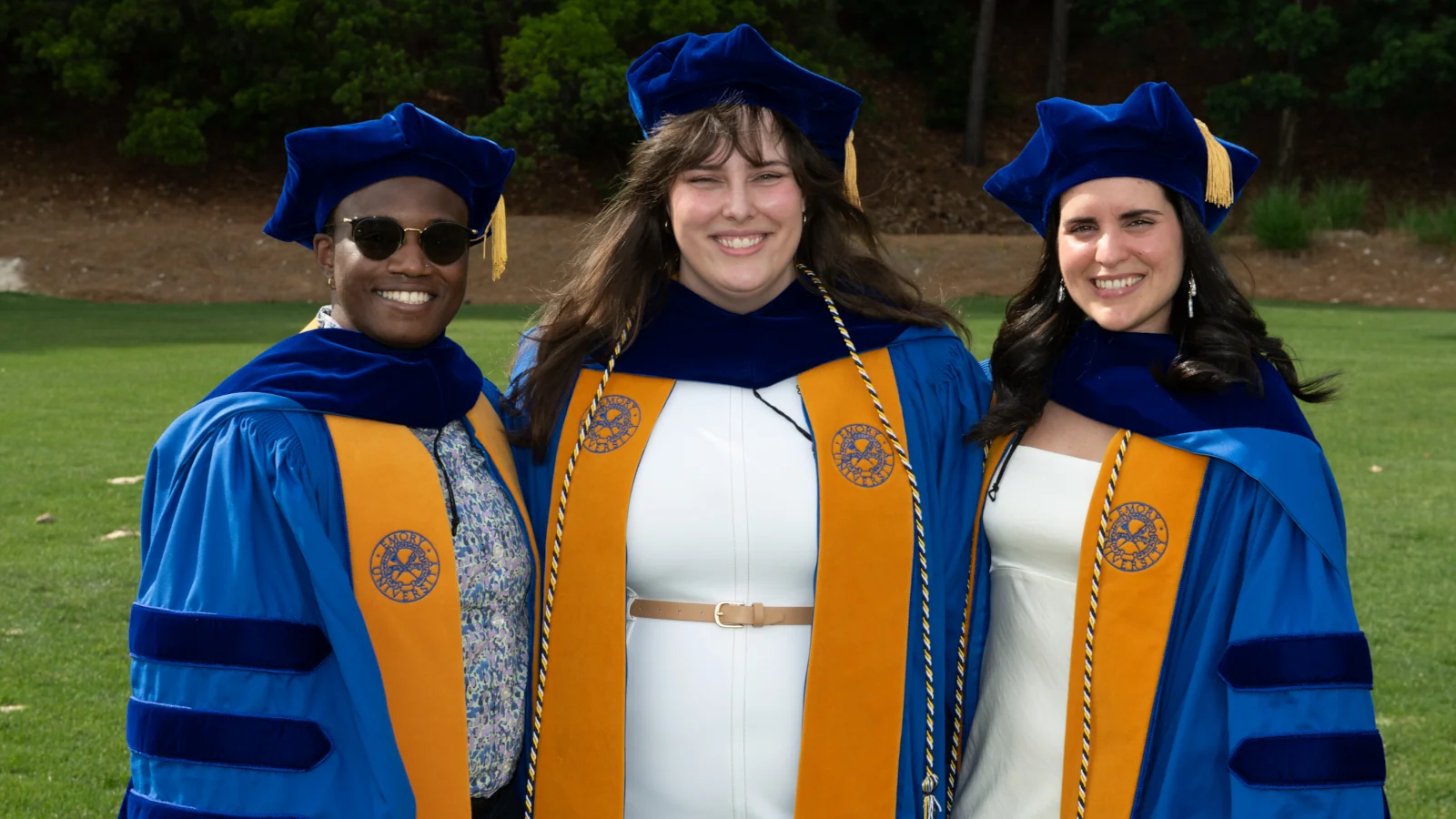Three doctoral graduates in academic regalia smile outdoors after a commencement ceremony.