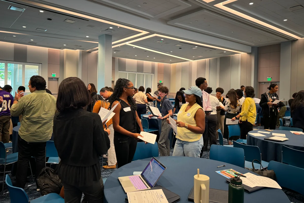Graduate students participate in a large group activity in a brightly lit conference space, holding papers and engaging in conversation.