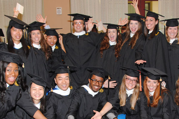 The photo includes 14 male and female graduates in black commencement regalia celebrating happily. 