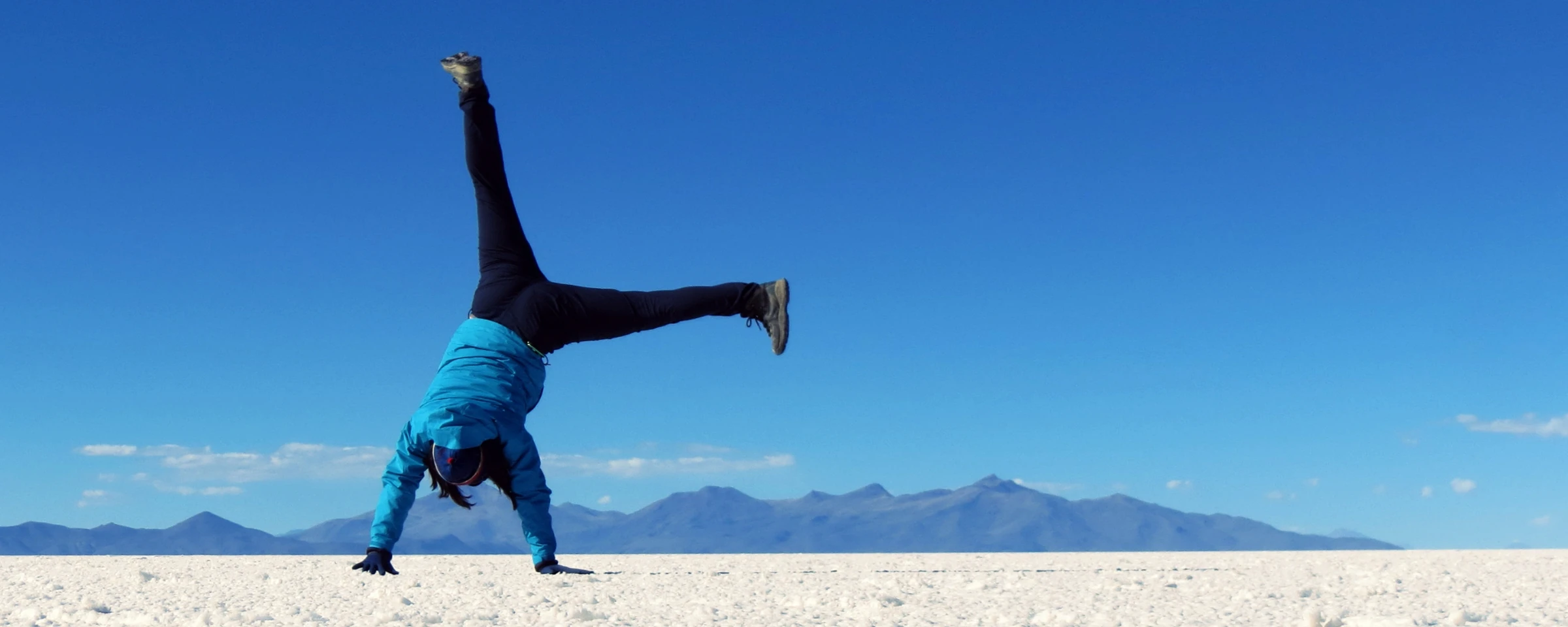 Student performing a one-handed handstand on the white salt flats of Bolivia with mountains in the background under a clear blue sky.